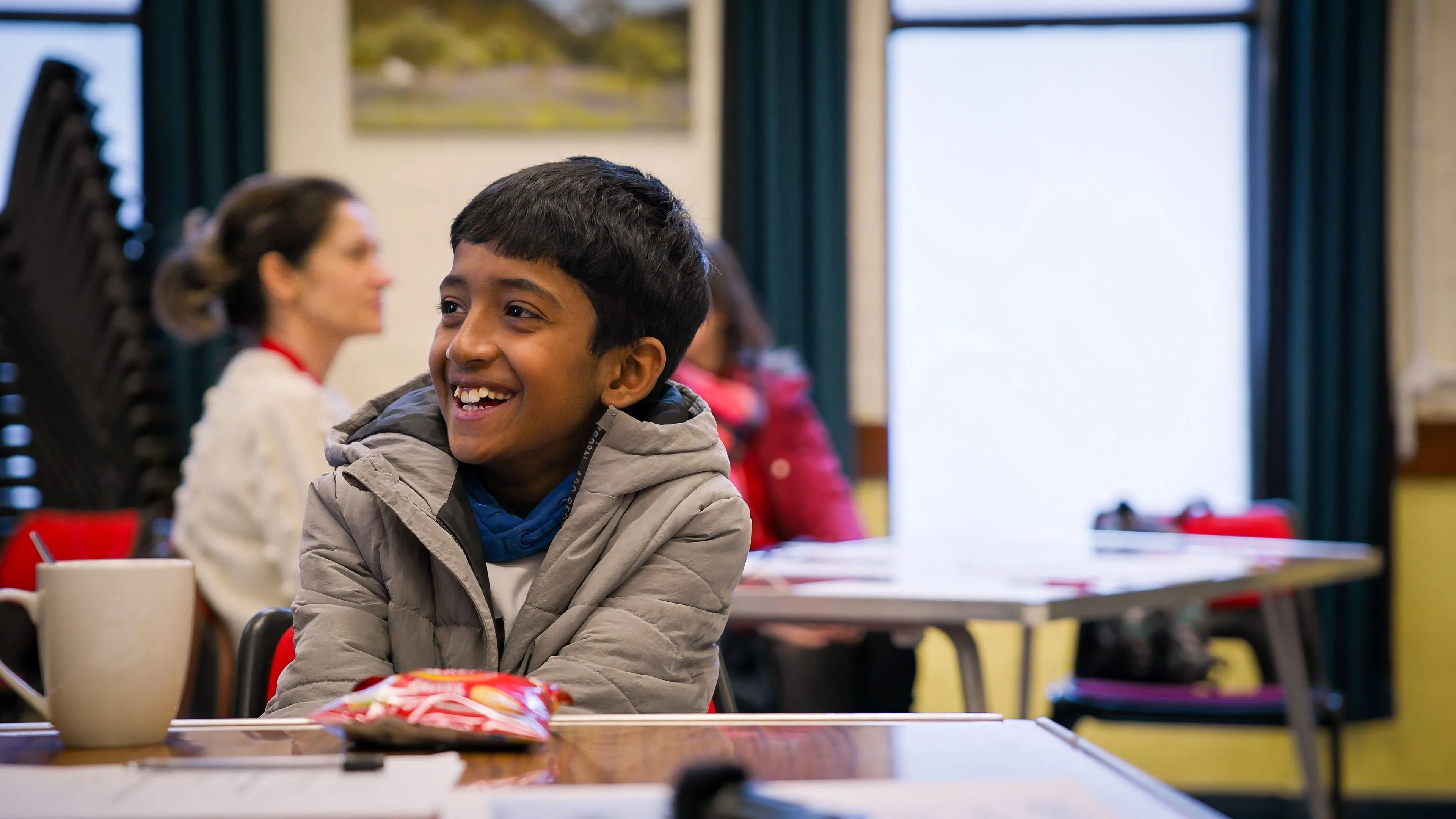 A young boy in a gray jacket smiles brightly while sitting at a table with a cup and snacks. People are blurred in the background, suggesting a lively setting.